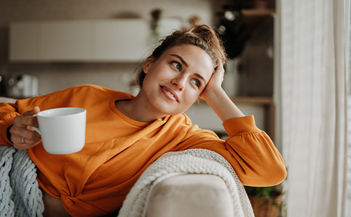Frau liegt auf dem Sofa und hält eine Tasse in der Hand. 