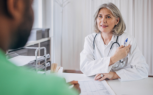 Eine Ärztin sitzt mit einem Patienten, den man nur von hinten sieht, an einem Schreibtisch und bespricht etwas.