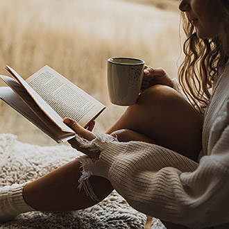 Person sitzt eingekuschelt und liest ein Buch neben einer Tasse Getränk