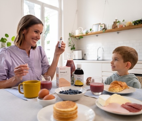 Una madre mostra al figlio una bottiglietta di Omega-3 durante la colazione; sulla tavola ci sono integratori, pancake e succo.