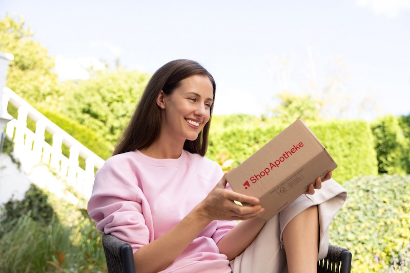 Eine lächelnde Frau sitzt im Garten und hält ein Paket von Shop Apotheke in der Hand.
