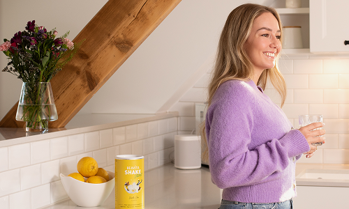 Eine lächelnde Frau in einem lilafarbenen Pullover steht in einer Küche und hält ein Glas Wasser in der Hand.