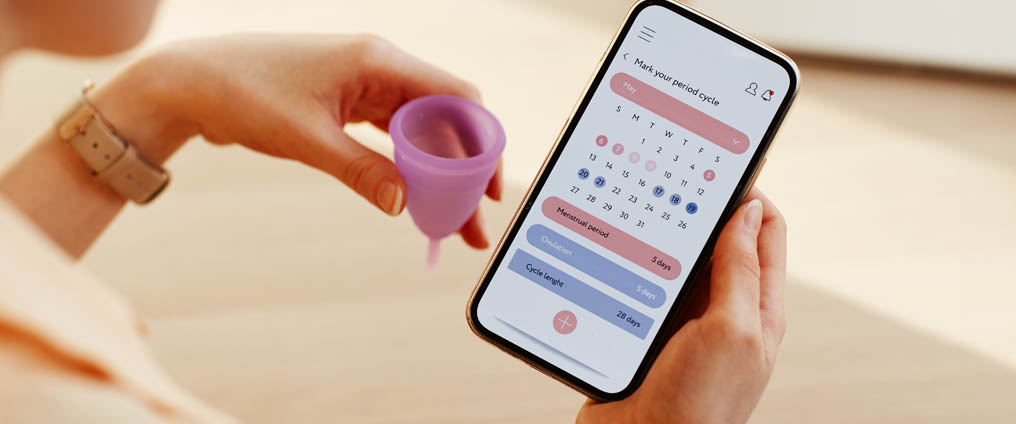 Eine Person hält eine violette Menstruationstasse in der Hand und schaut auf ein Smartphone mit geöffneter Perioden-Tracking-App.