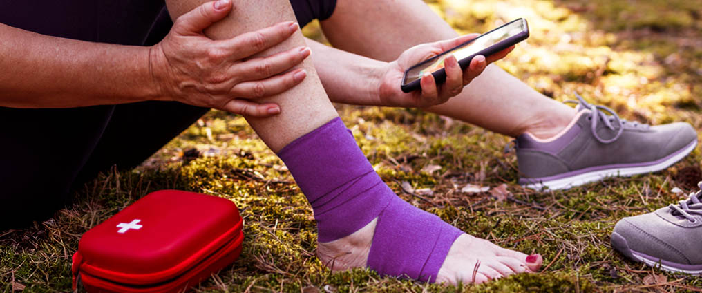Eine Person sitzt im Freien, hat den Knöchel mit einer lila Bandage verbunden und hält ein Smartphone in der Hand, daneben liegt ein rotes Erste-Hilfe-Set.