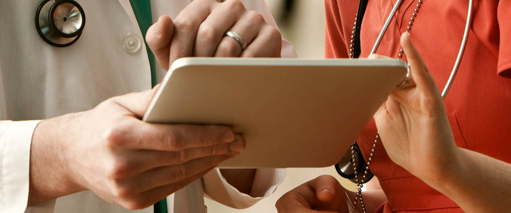 Arzt oder Ärztin hält Tablet in der Hand und schaut mit einer weiteren Person darauf.