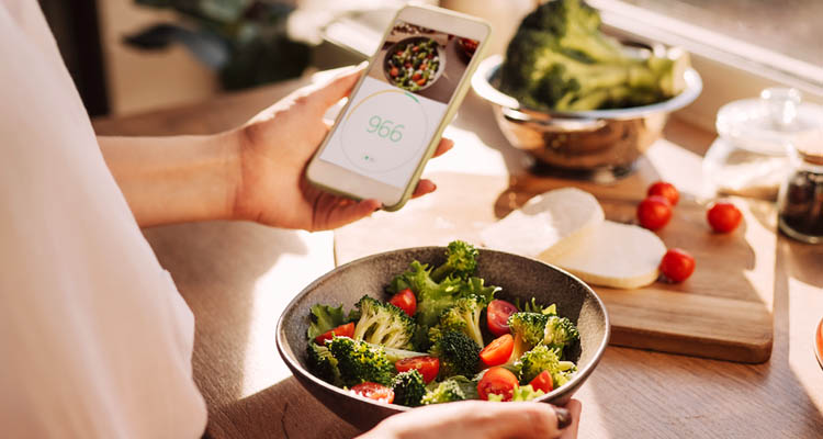 Person hält Smartphone mit App-Anzeige, davor Schüssel mit Salat, Tomaten und weitere Zutaten auf Küchentheke.
