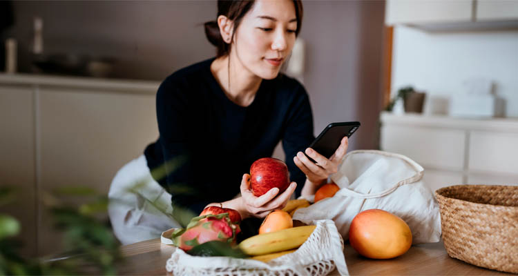 Eine Frau sitzt in der Küche, hält ein rotes Apfel in der Hand und schaut auf ihr Smartphone, vor ihr liegen verschiedene frische Früchte.