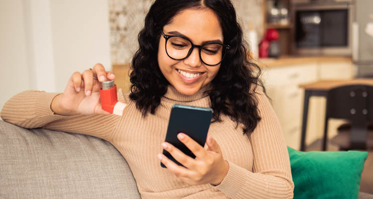 Eine Frau mit Brille sitzt auf dem Sofa, hält einen Asthma-Inhalator in der einen Hand und schaut lächelnd auf ihr Smartphone.