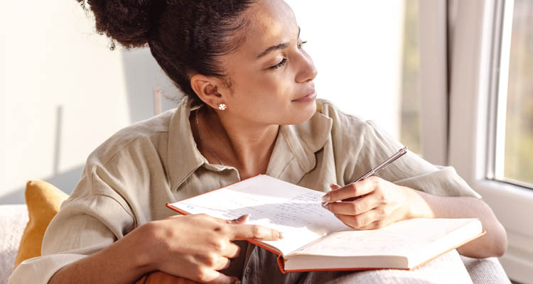 Frau mit hochgestecktem Haar sitzt an einem Tisch neben einem Fenster und hält ein aufgeschlagenes Buch in der Hand.