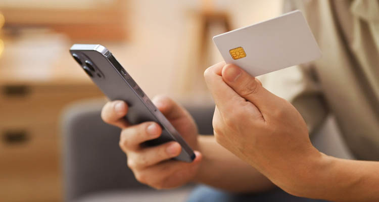 Hand hält eine weiße Kreditkarte mit Chip und ein Smartphone mit schwarzem Gehäuse in einem Wohnzimmer.