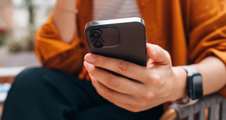 Nahaufnahme einer Hand, die ein Smartphone hält; die Person trägt ein orangefarbenes Oberteil und eine Smartwatch..