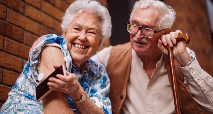 Eine ältere Frau zeigt lachend einen am Oberarm befestigten Gesundheitssensor, neben ihr steht ihr Partner mit Gehstock.