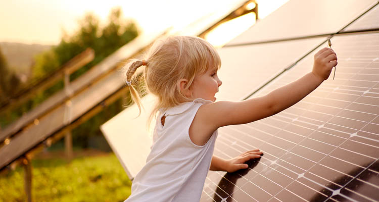 Ein blondes Kind im weißen Shirt berührt ein Solarpanel in der Abendsonne.