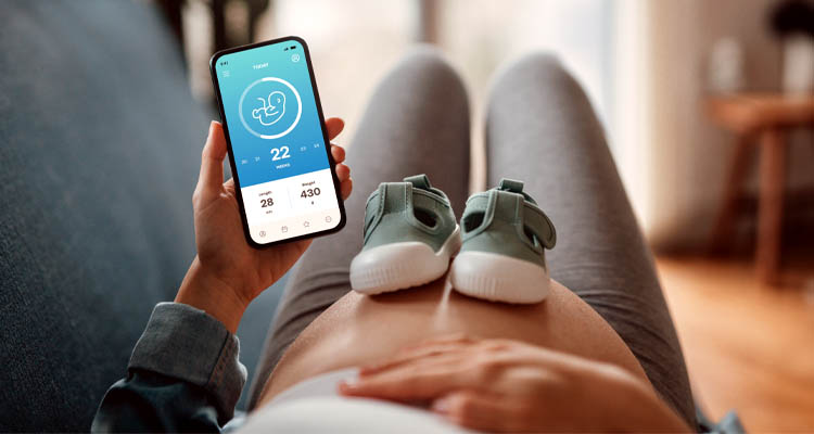 Eine schwangere Person liegt auf dem Sofa, auf dem Bauch stehen Babyschuhe, in der Hand ein Smartphone mit Schwangerschafts-App.