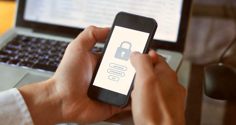 Eine Person hält ein Smartphone mit geöffneter Login-Maske mit Schloss-Symbol in der Hand, im Hintergrund ein Laptop und eine Tasse Kaffee.