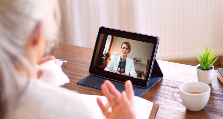 Tablet auf einem Tisch zeigt eine Videokonferenz mit einer Person in weißem Arztkittel, daneben eine Tasse Kaffee und eine grüne Pflanze.
