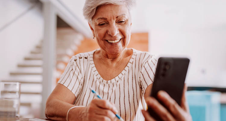 Ältere Frau mit weißem Haar und gestreiftem T-Shirt sitzt am Tisch, hält Smartphone in der linken Hand und schreibt mit einem Stift auf Papier.