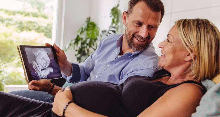 Ein Mann und eine schwangere Frau sitzen auf dem Sofa und schauen gemeinsam ein Ultraschallbild auf einem Tablet an.