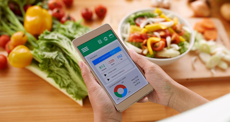Hand hält Smartphone mit Gesundheits-App, die Schritte, Kalorien und Aktivitätszeit anzeigt, im Hintergrund frisches Gemüse und Salat auf Holzbrett.