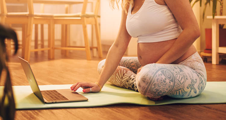 Schwangere Frau in weißem Top und gemusterten Leggings sitzt auf Yogamatte und nutzt Laptop in hellem Raum mit Holzstühlen im Hintergrund.