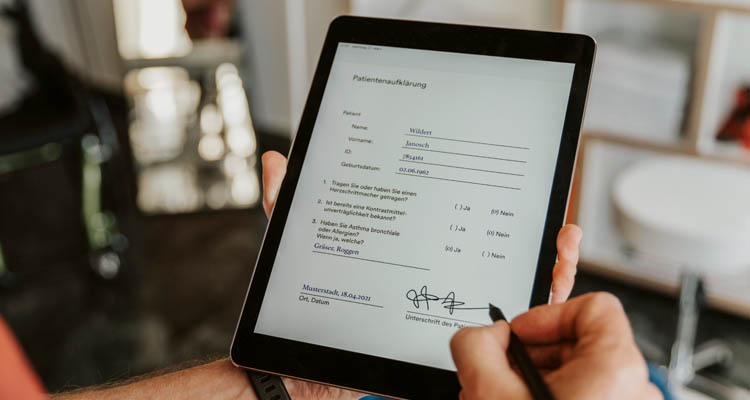 Eine Person hält ein Tablet und füllt darauf ein digitales Patientenformular mit Unterschrift aus.