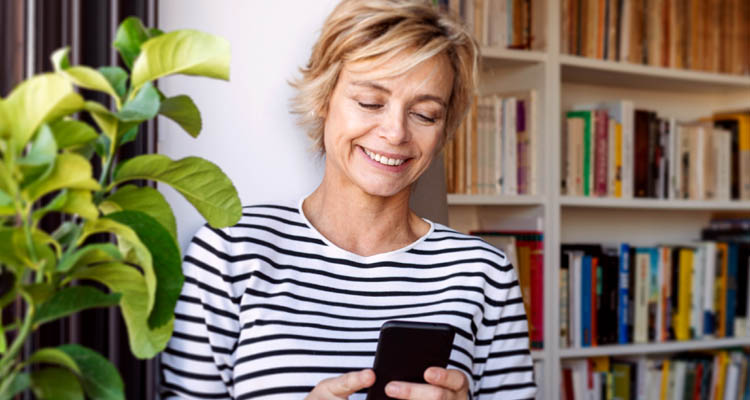 Eine lächelnde Frau im gestreiften Shirt steht vor einem Bücherregal und tippt auf ihr Smartphone.