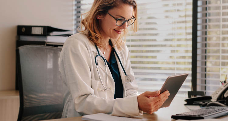 Eine Ärztin im weißen Kittel mit Stethoskop betrachtet ein Tablet an ihrem Schreibtisch.