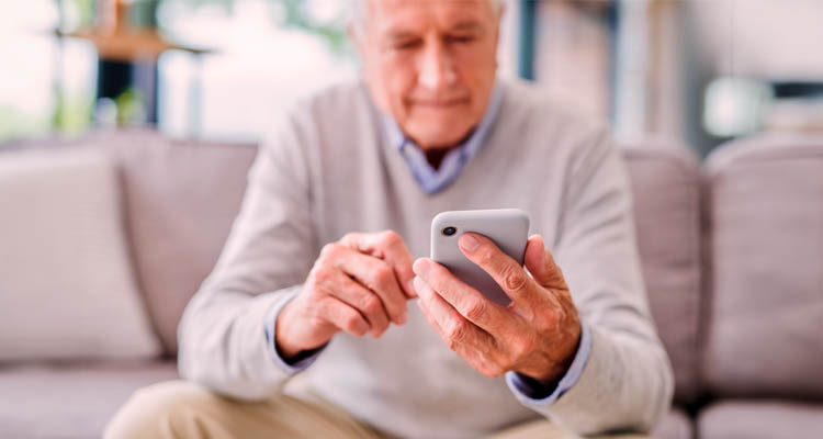 Ältere Person sitzt auf einem Sofa und hält ein weißes Smartphone in der rechten Hand.