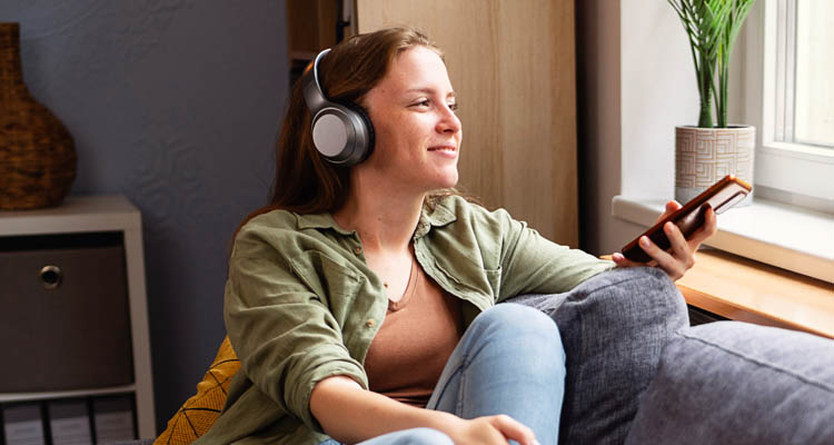 Eine junge Frau sitzt entspannt am Fenster, trägt große Kopfhörer und hält ein Smartphone in der Hand.
