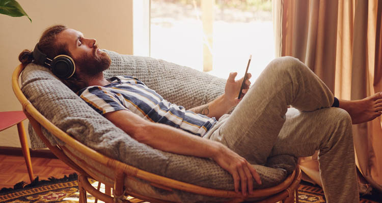 Ein Mann mit Kopfhörern sitzt entspannt in einem Sessel, hält sein Smartphone locker in der Hand.