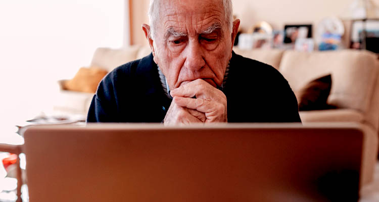 Älterer Mann sitzt vor einem Laptop, Hände vor dem Mund gefaltet, im Wohnzimmer mit Sofa und Kissen im Hintergrund.