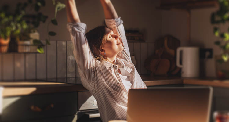 Person sitzt vor einem Laptop und streckt die Arme nach oben, trägt ein gestreiftes Hemd, im Hintergrund Küchenutensilien und Pflanzen.