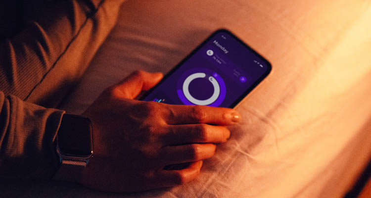 Eine Person liegt im Bett und hält ein Smartphone mit geöffneter Schlaftracking-App, neben ihr eine Smartwatch.