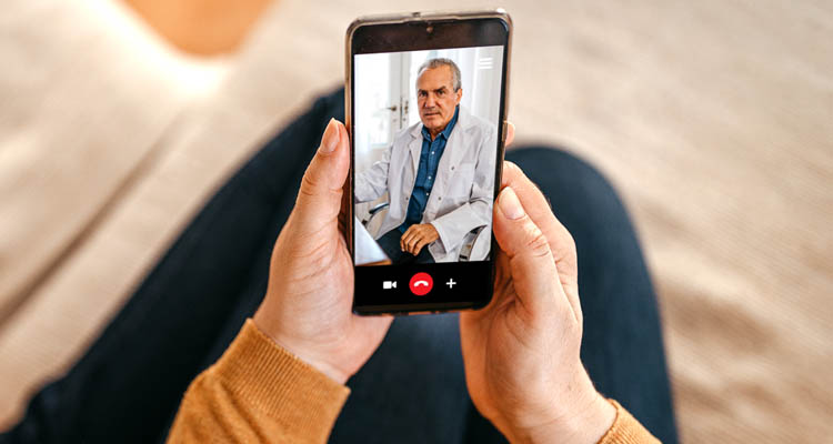 Hände halten ein Smartphone mit einem Videoanruf, darauf ein sitzender Arzt in weißem Kittel und Hemd zu sehen.