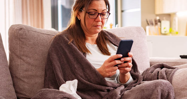 Person sitzt auf einem Sofa, eingewickelt in eine braune Decke, hält ein Smartphone in beiden Händen, neben ihr liegt ein zerknülltes Taschentuch.