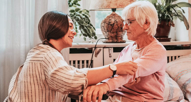 Junge Frau misst Blutdruck einer älteren Frau mit Manschette am Oberarm in einem Wohnzimmer mit Pflanzen und Vasen.