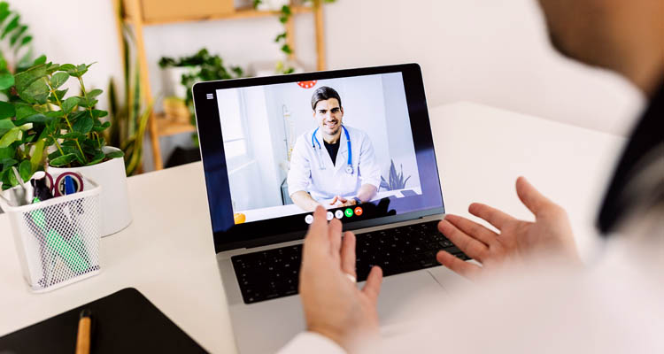 Person sitzt vor einem Laptop und führt eine Videokonferenz mit einer Ärztin in weißem Kittel und Stethoskop durch.