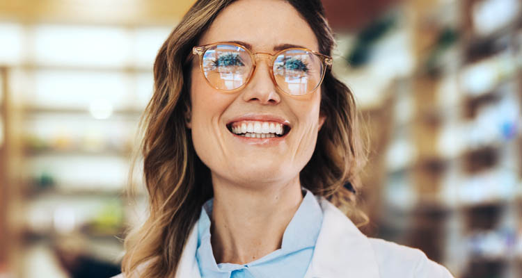 Eine Frau mit Brille und weißem Kittel lächelt in einer Apotheke, im Hintergrund sind Regale mit Medikamenten zu sehen.