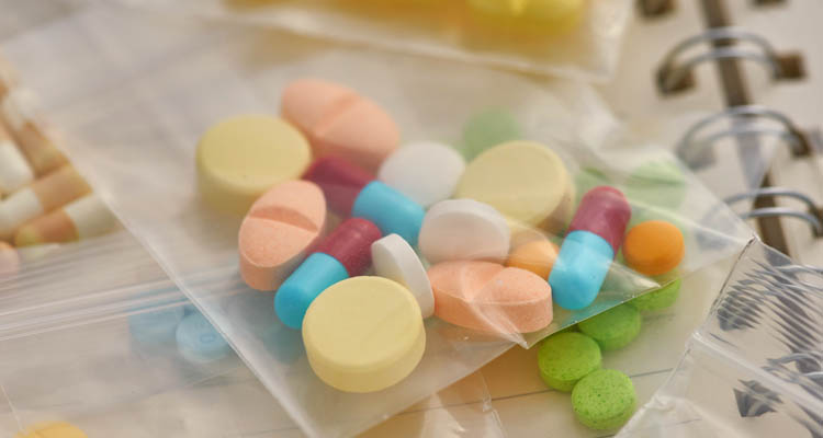 Mehrere bunte Tabletten und Kapseln in durchsichtigen Plastikbeuteln auf einem Tisch neben einem Spiralnotizbuch.