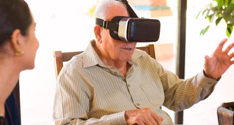 Älterer Mann sitzt auf einem Stuhl und trägt eine VR-Brille, hebt die rechte Hand, eine Frau sitzt ihm gegenüber.