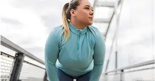 Frau in hellblauer Kapuzenjacke, die auf einer Metallbrücke leicht nach vorne gebeugt steht und mit den Händen auf den Knien verschnauft.