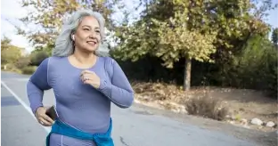 Ältere Frau mit grauen Haaren in lila Sportshirt, die auf einem von Bäumen gesäumten Weg joggt und ein Smartphone in der Hand hält.
