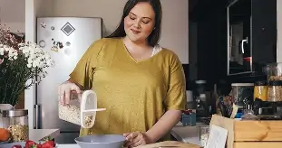 Frau in gelbem T-Shirt in einer Küche, die Frühstücksflocken aus einem Vorratsbehälter in eine Schüssel gießt.