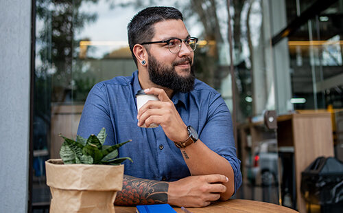 Junger kräftiger Mann sitzt draußen in einem Café und trinkt aus einer Tasse.