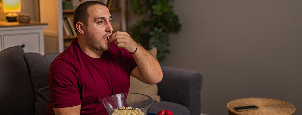Ein kräftiger Mann sitzt auf der Couch und isst Popcorn.