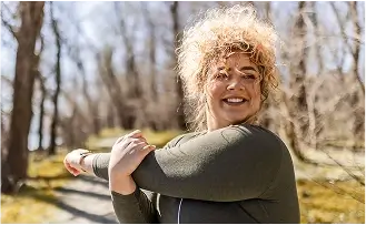 Frau in Sportbekleidung, die im Wald auf einem Wanderweg ihre Arme dehnt und dabei lächelt.