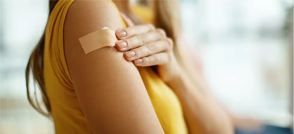 Frau mit gelbem Oberteil berührt vorsichtig das Pflaster auf ihrem Oberarm nach einer Impfung.