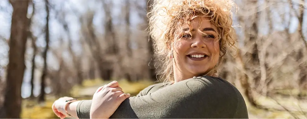 Frau in Sportbekleidung, die im Wald auf einem Wanderweg ihre Arme dehnt und dabei lächelt.