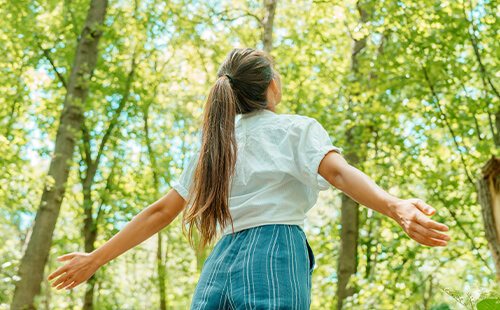 Eine Frau steht mit geöffneten Armen im Wald und atmet die frische Luft ein. 