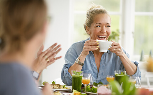 Zwei Frauen sitzen gemeinsam am Tisch, die eine Frau trinkt aus einer Tasse und lacht. 